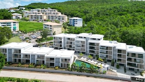 A modern hotel complex with multiple buildings, a swimming pool, and lush greenery in the background.