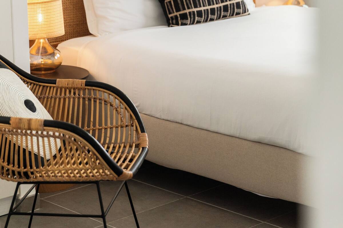 Un lit bien fait, avec des oreillers blancs et noirs, une chaise en osier et une lampe de chevet.