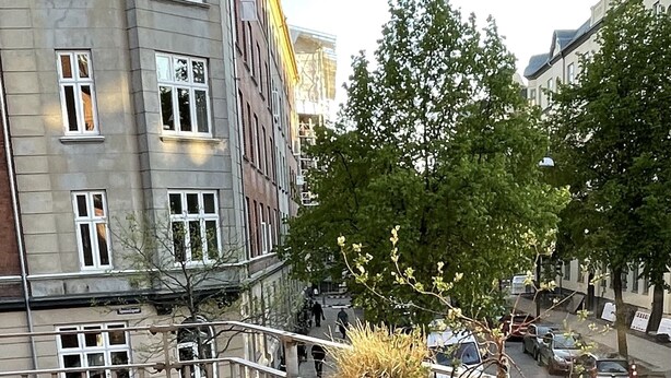 A balcony with a table and chairs, potted plants, and a view of a city street with buildings and cars.