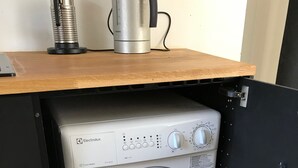 A compact, modern washing machine with a control panel, placed under a wooden shelf.