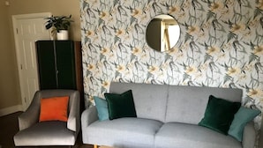 Un salon avec un canapé gris, des coussins turquoise et verts, une table basse en bois et un papier peint à motifs floraux.