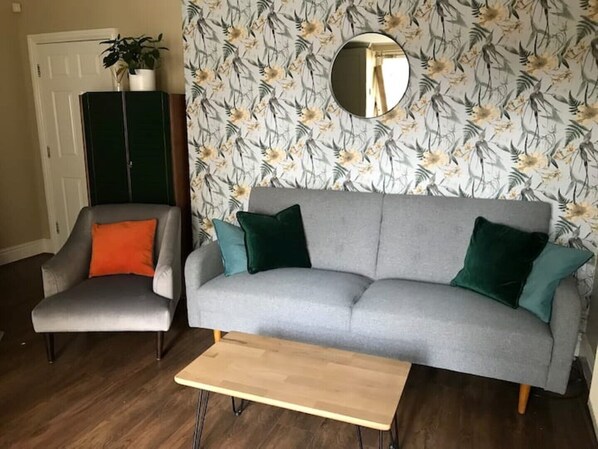 Un salón con un sofá gris, cojines verde azulado y verde, una mesa de centro de madera y un papel tapiz floral.