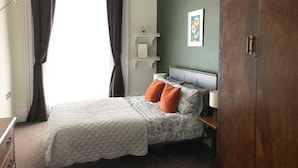 Une chambre à coucher comprenant un lit, une armoire, une table de chevet, une commode, une fenêtre avec des rideaux et une étagère fixée au mur.