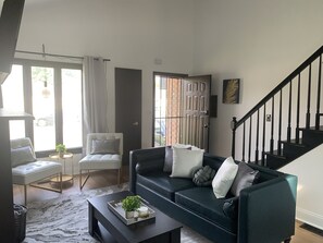 A living room with a dark green sofa, a white armchair, and a black coffee table. There is a staircase with black railings and a door with a metal gate.
