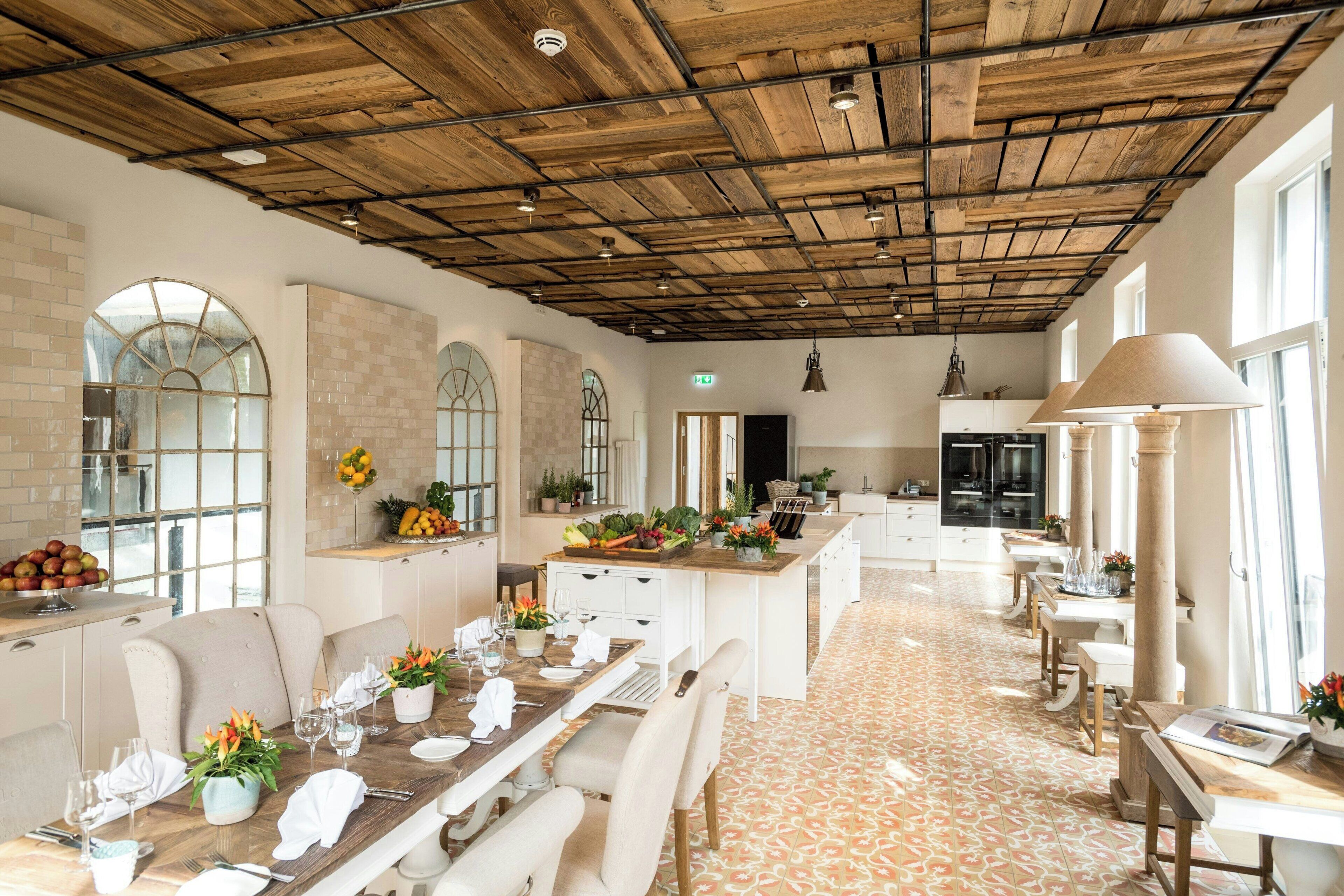 A spacious dining area with a long wooden table set for a meal, surrounded by white chairs. A kitchen area is visible in the background with modern appliances and a large window providing natural light.