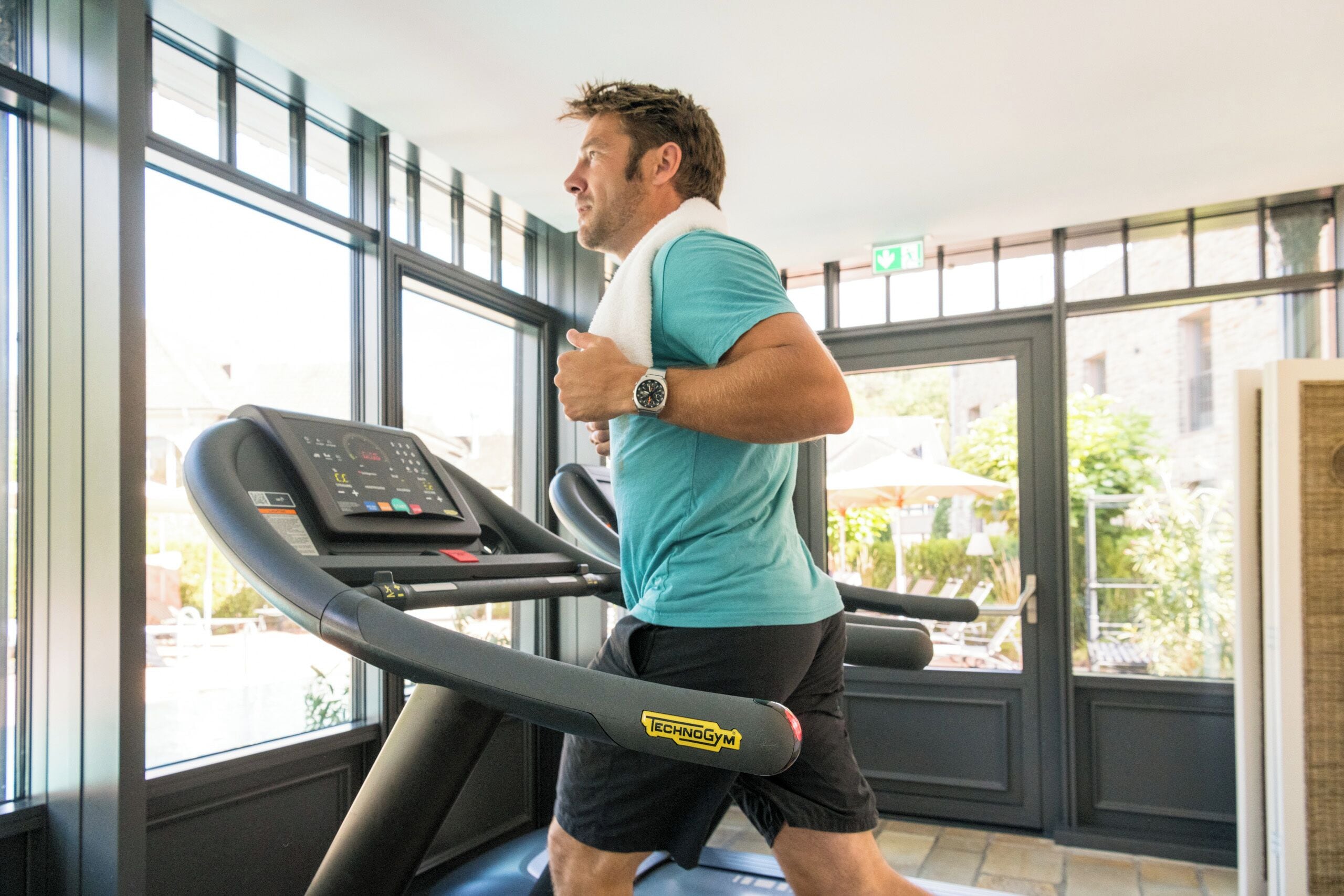 Un uomo corre su un tapis roulant in palestra, con ampie finestre e vista sull'esterno.