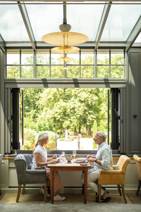 A dining area with a table and chairs, large windows with a view of trees, and pendant lighting. - Bollants SPA im Park, Autograph Collection (Bad Sobernheim)