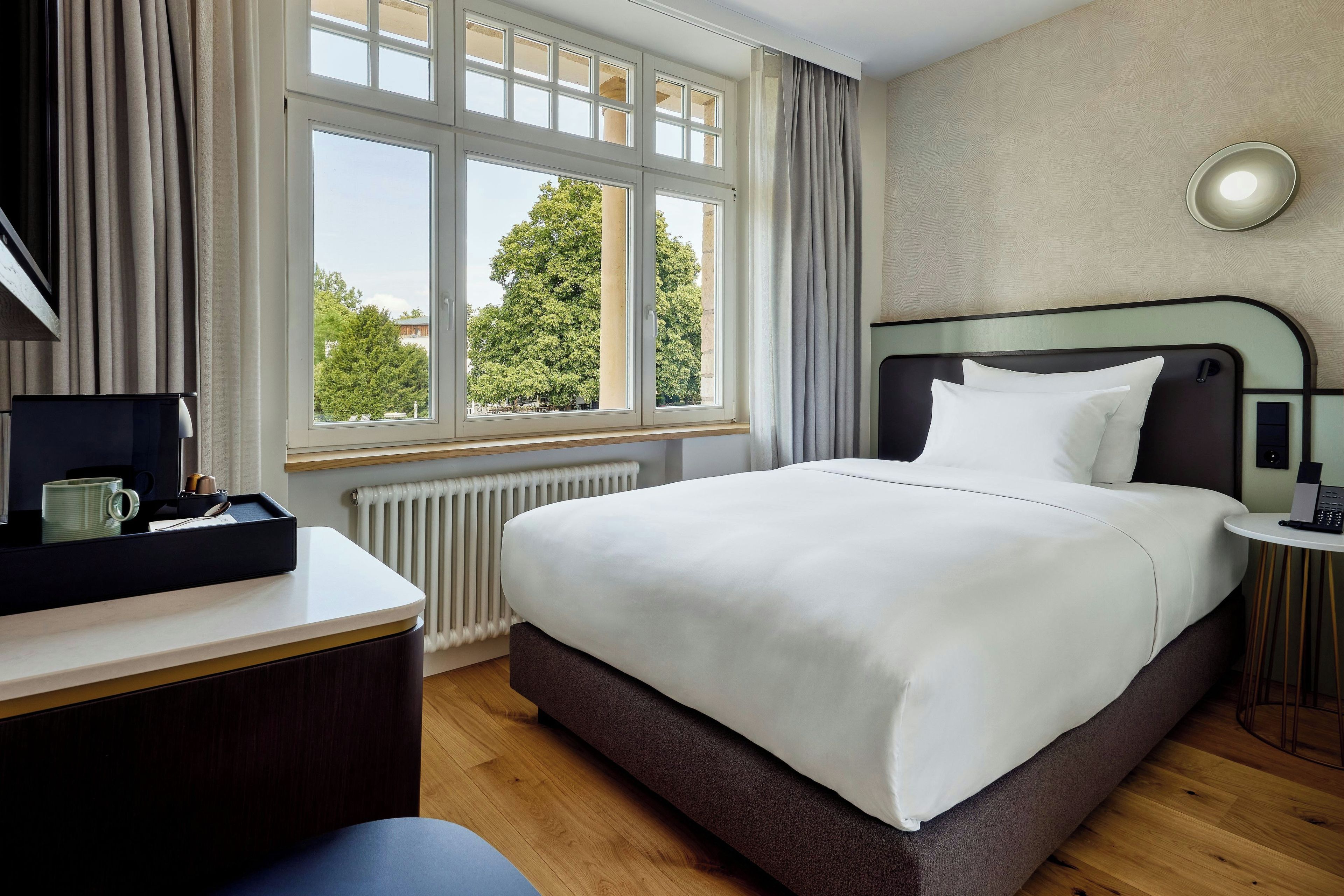 Habitación de hotel con cama, escritorio, silla, ventana con cortinas y radiador.