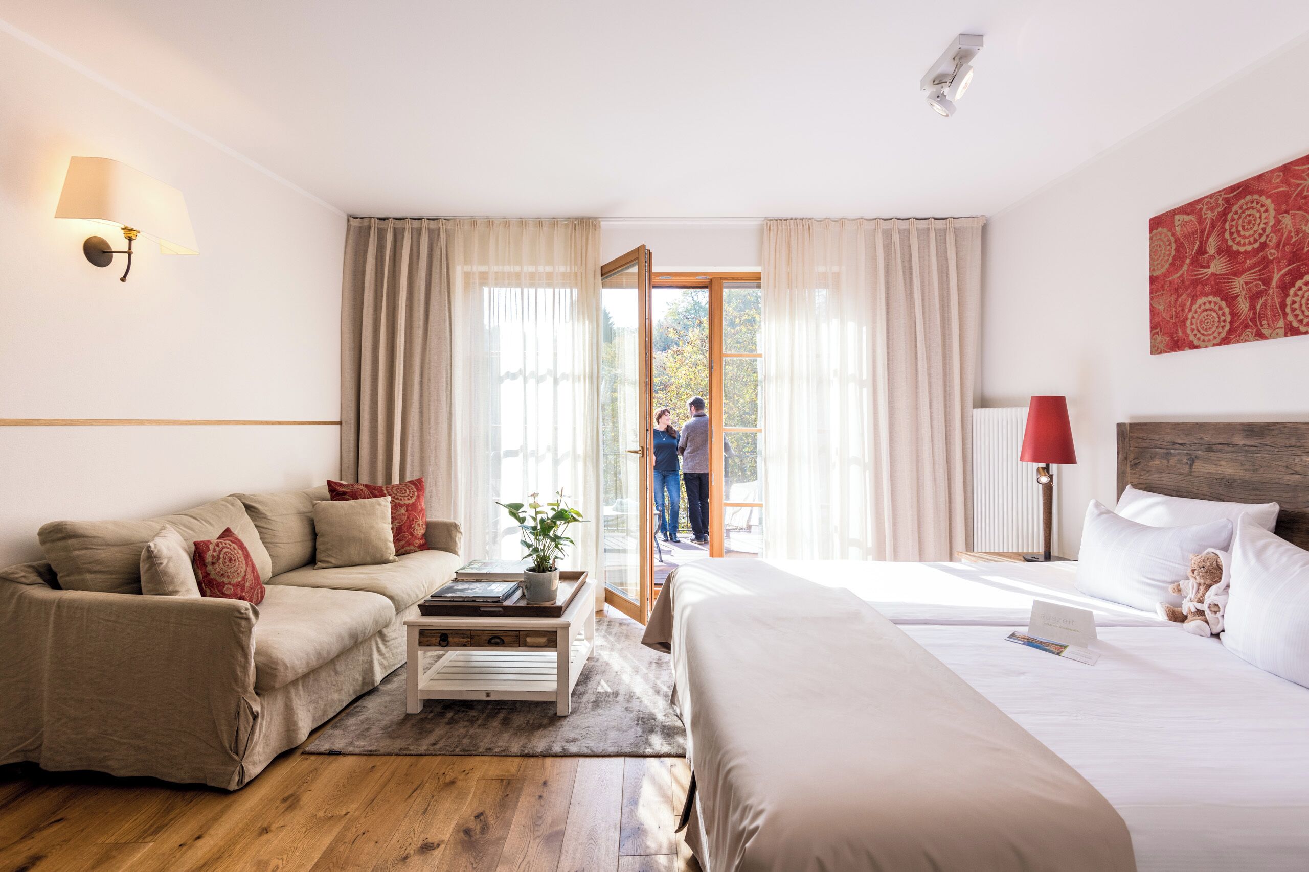 A hotel room with a sofa, a bed, a bedside table, a lamp, and a view of the outside.
