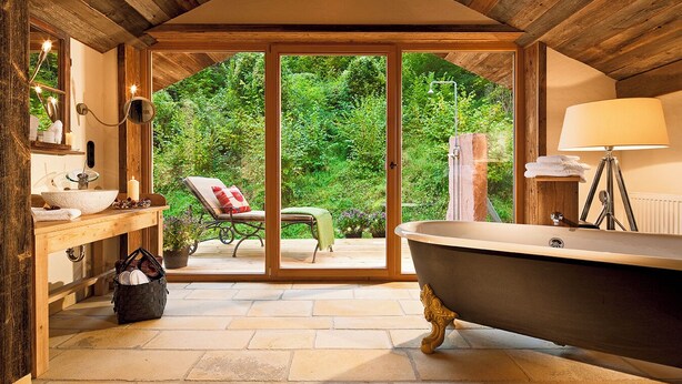 Un baño con bañera, lavamanos y una silla junto a un ventanal corredizo que da a un jardín.