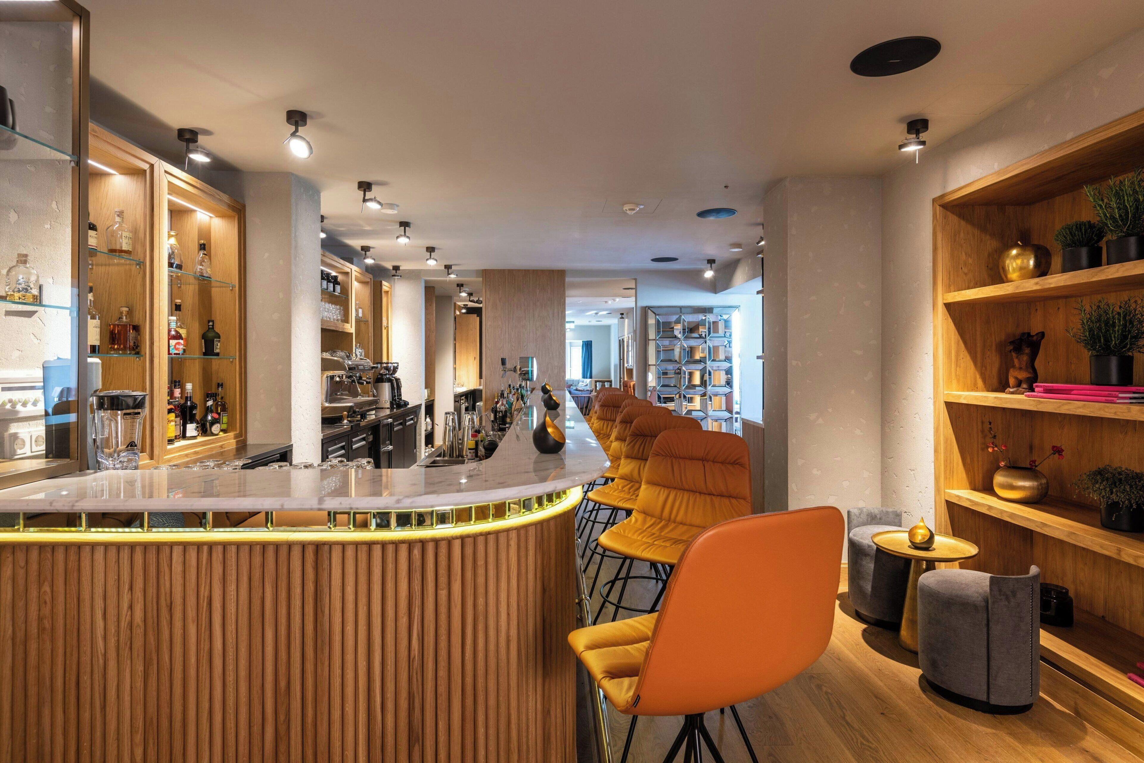 Un área de bar moderna con un mostrador de madera, taburetes y estantes con botellas.