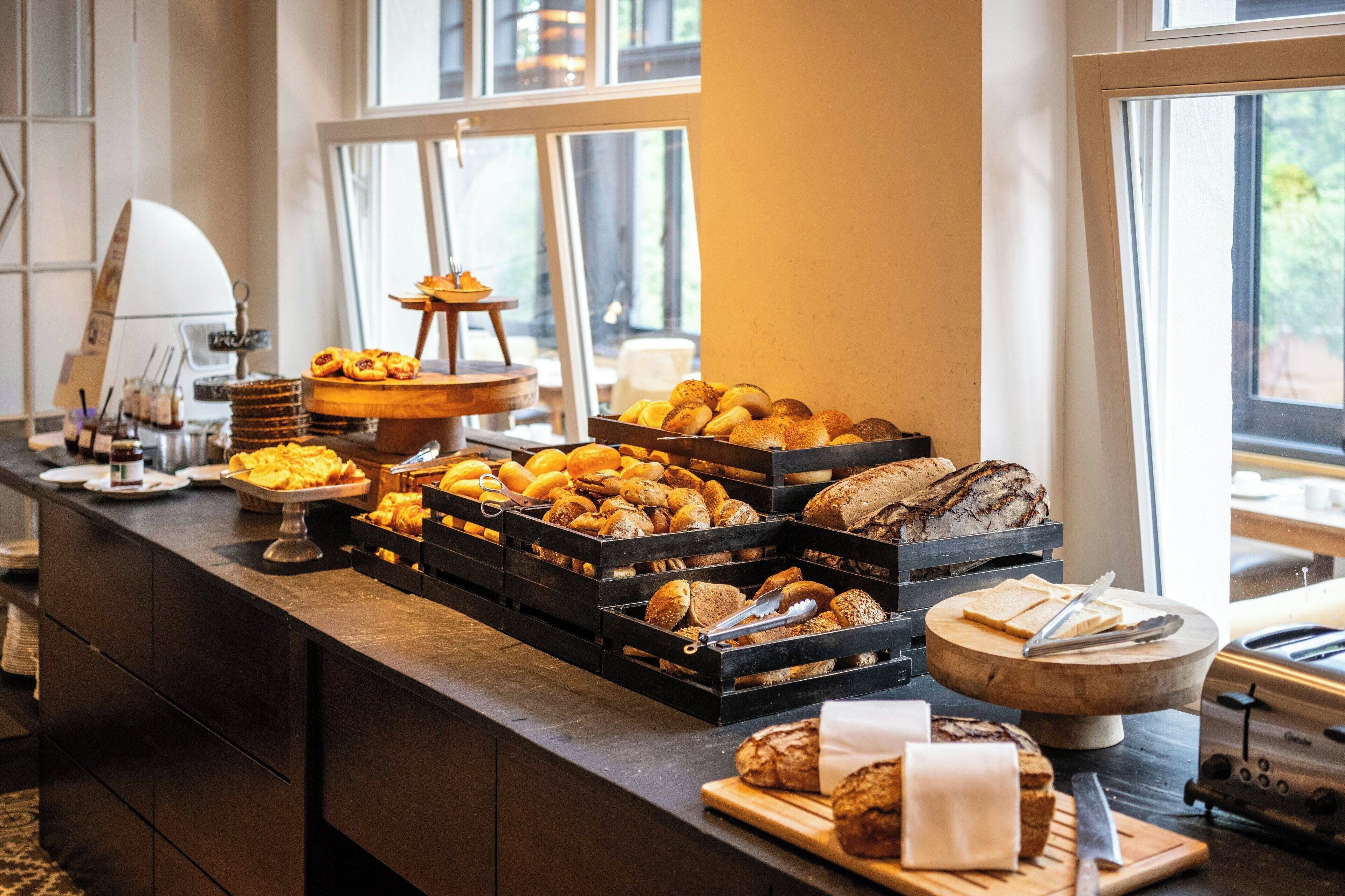 Ein Buffet mit verschiedenen Brotsorten und Gebäck, eine Kaffeestation und Blick ins Freie durch große Fenster.