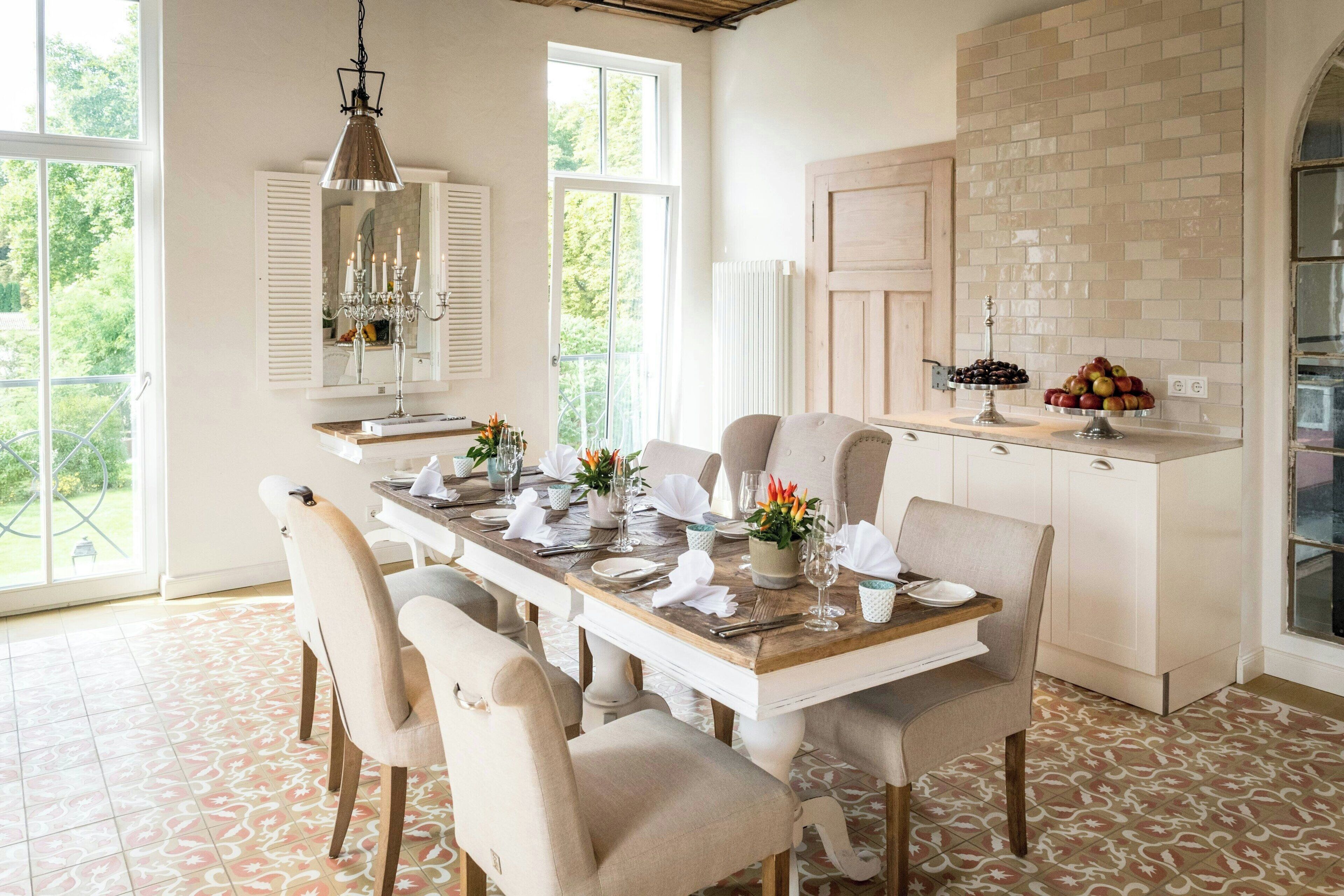 A dining area with a wooden table set for six, surrounded by upholstered chairs. A chandelier hangs above, and a buffet with fruits and a cake is visible on the counter.