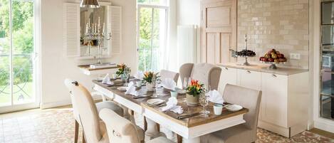 Un comedor con una mesa de madera dispuesta para seis personas, rodeada de sillas tapizadas. Una araña cuelga encima, y se ve un bufé con frutas y un pastel en la encimera.