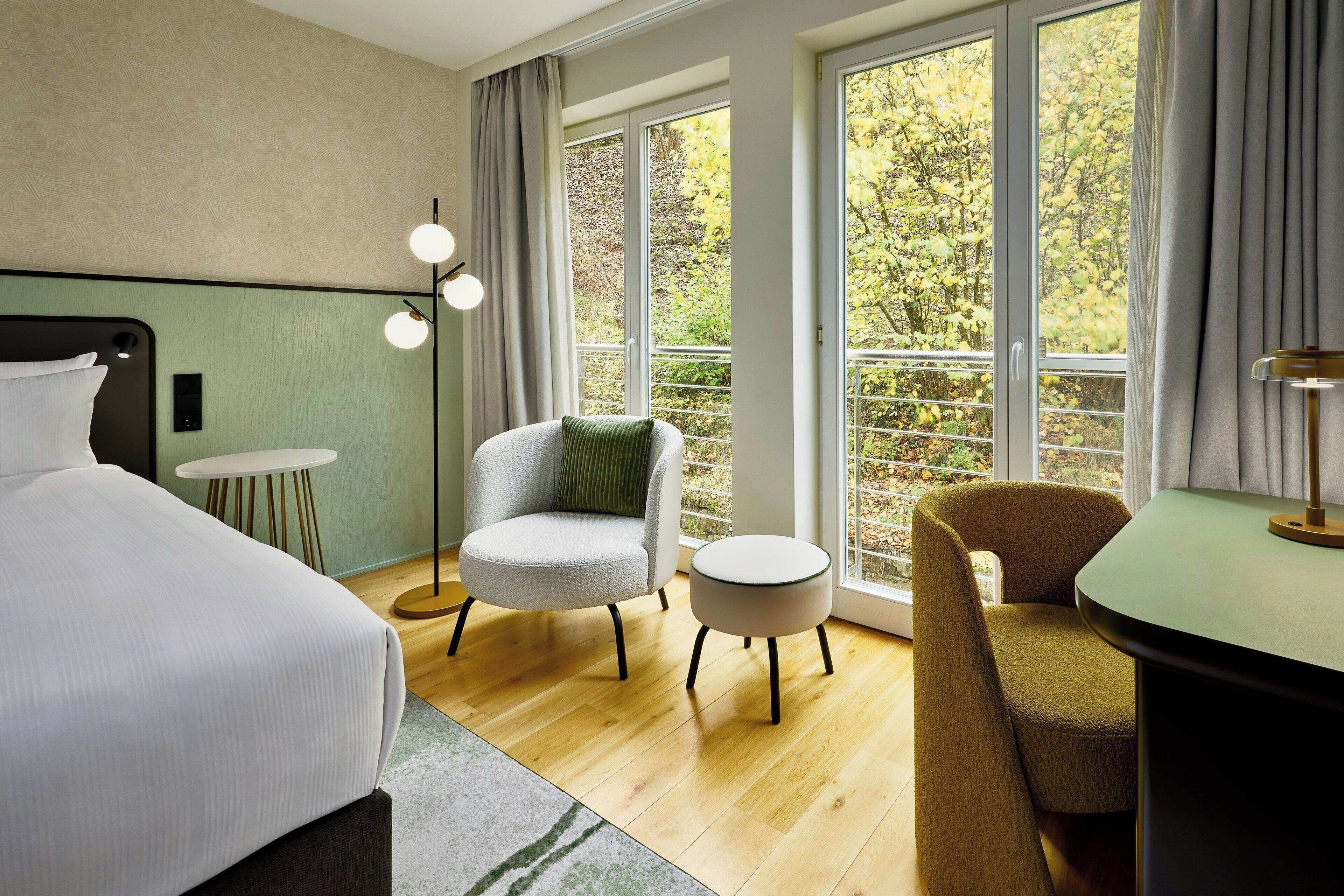 A modern hotel room with a bed, two chairs, a small table, and a large window offering a view of trees.