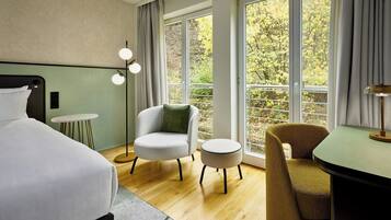 A modern hotel room with a bed, two chairs, a small table, and a large window offering a view of trees.