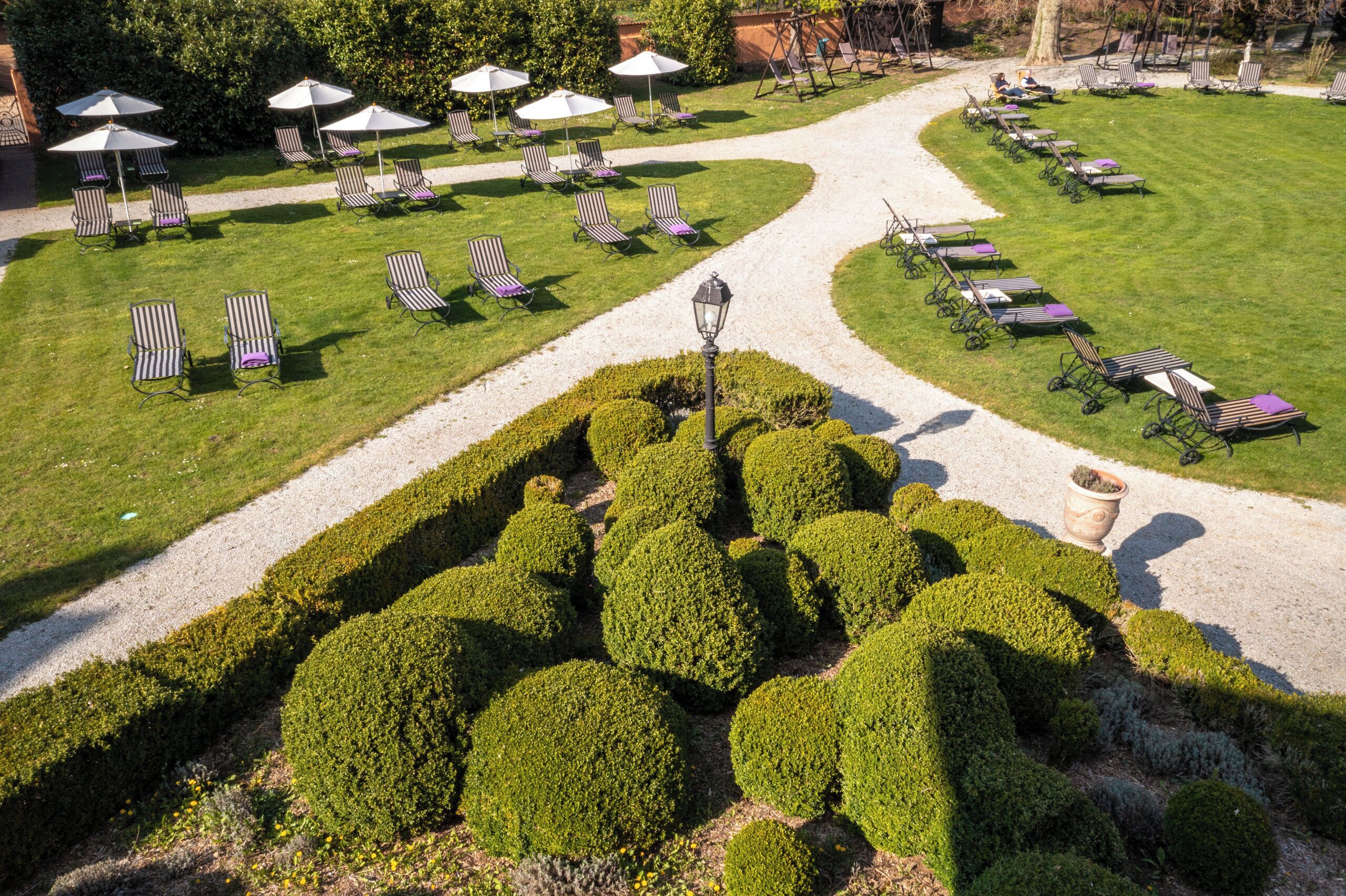 A well-maintained garden with neatly trimmed hedges, a gravel path, and rows of sun loungers with umbrellas.