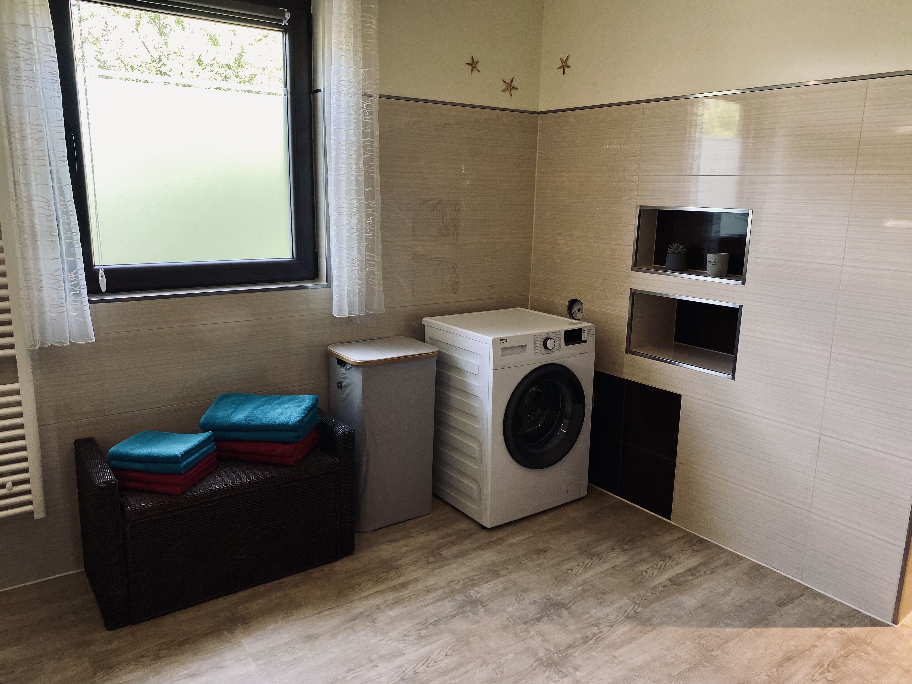 A compact laundry room with a washing machine, a small shelf, and a wicker bench with folded clothes.