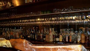 Un bar avec un grand comptoir en bois où sont exposés des produits de viande emballés et diverses bouteilles d’alcool.