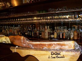 Un bar con una amplia barra de madera que exhibe un producto cárnico envuelto y diversas botellas de alcohol.