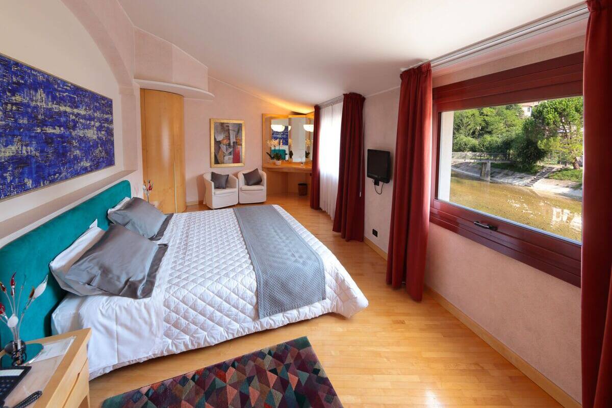 Ett modernt hotellrum med en stor säng, ett skrivbord med en dator, en stol, en TV och ett fönster med utsikt över en vattenyta.
