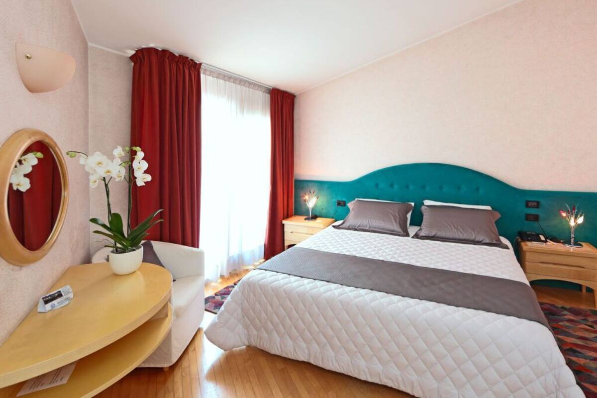 Ett hotellrum med en säng, sängbord, ett skrivbord, en stol, en spegel och en krukväxt.