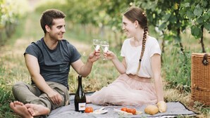 Un couple est assis sur une nappe à pique-nique dans un vignoble, tenant des verres de vin blanc. Une bouteille et des aliments sont posés sur la nappe.