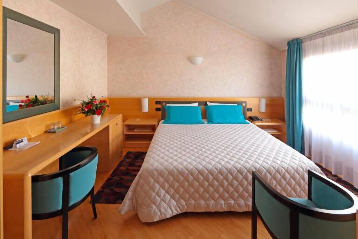 Ett hotellrum med en stor säng, ett skrivbord, en stol, en spegel och ett fönster med gardiner.