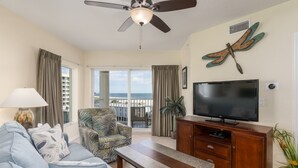 A living room with a ceiling fan, a television, a patterned armchair, a sofa, a wooden coffee table, and a view of the ocean through large windows. - 607S-Penthouse-Gulf View, Top Floor, 2/2 on Beach (Treasure Island)