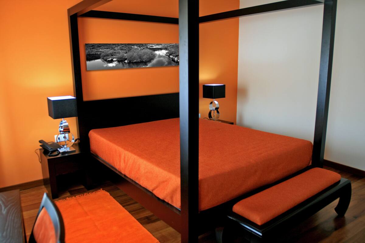 Een hemelbed met een oranje sprei, een nachtkastje met een lamp, en een zwart-wit foto in een lijst aan de muur.