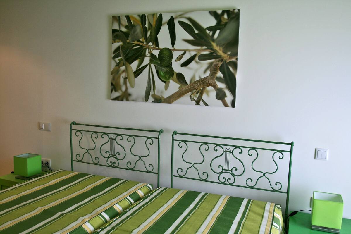 Een slaapkamer met twee bedden, een bedsprei met groene strepen, een kunstwerk van bladeren aan de muur, en een nachtkastje in het groen met een lamp.