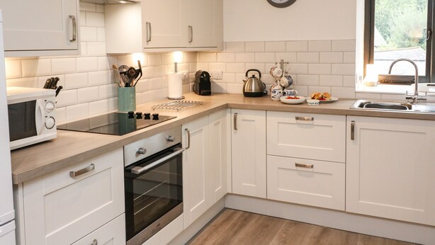 A modern kitchen with white cabinets, a built-in oven, a kettle, and a clock on the wall. - Primrose at Stancombe Manor, Nr Kingsbridge (Kingsbridge)