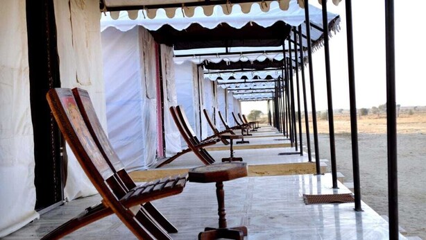 A row of wooden lounge chairs and a small table under a covered area with white curtains. - Shivas Heritage and Sand Dunes (Jaisalmer)