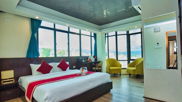 A hotel room with a large bed, a desk, and a chair. There are large windows with curtains, and a view of the city. - Hotel Grand View (Kathmandu)