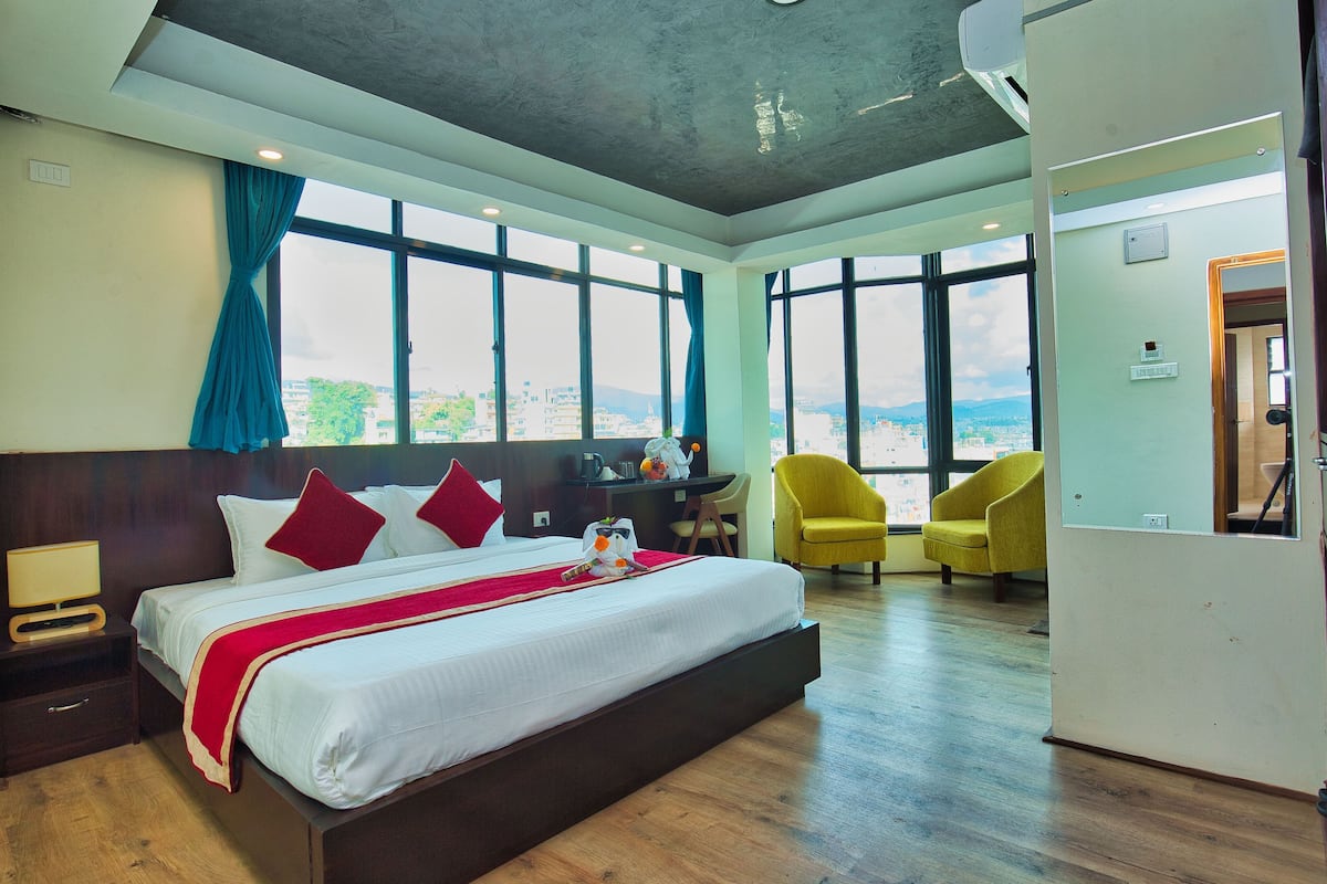 A hotel room with a large bed, a desk, and a chair. There are large windows with curtains, and a view of the city.