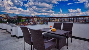 A rooftop terrace with wicker furniture and a table, overlooking a cityscape with mountains in the background. - Hotel Grand View (Kathmandu)