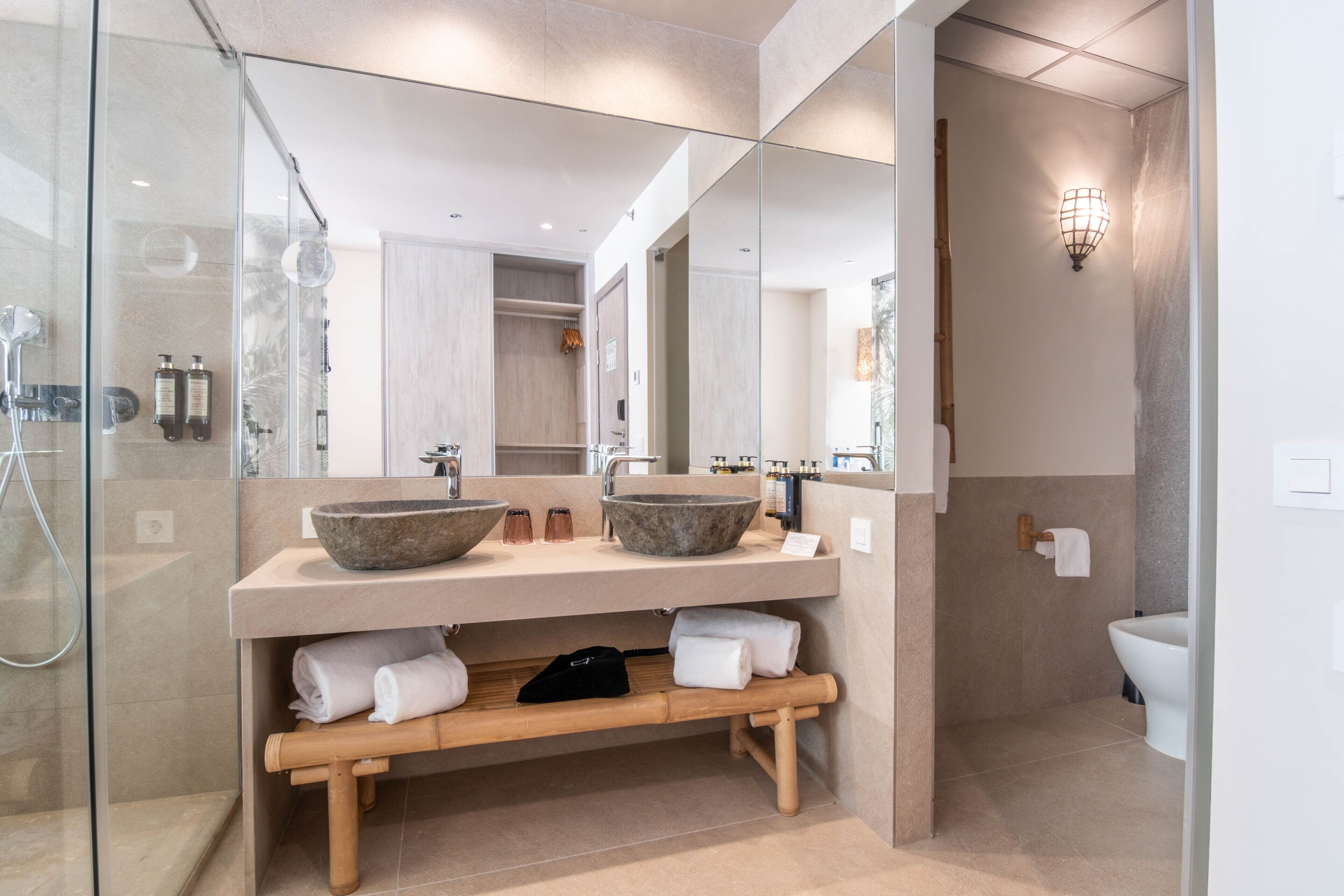 Un baño moderno con un espejo grande, dos lavamanos de piedra, una zona de ducha y un banco de madera.