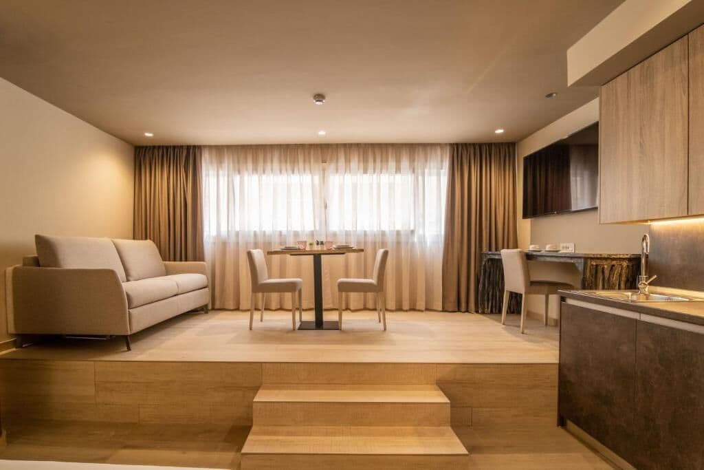 Una sala de estar moderna con un sofá, una mesa de comedor con sillas, una zona de cocina con un fregadero y un amplio ventanal con cortinas.