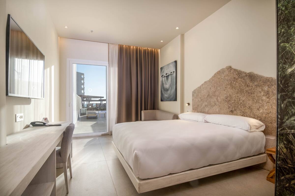 A modern hotel room with a large bed, a desk, and a chair. There is a view of the outside through a glass door.