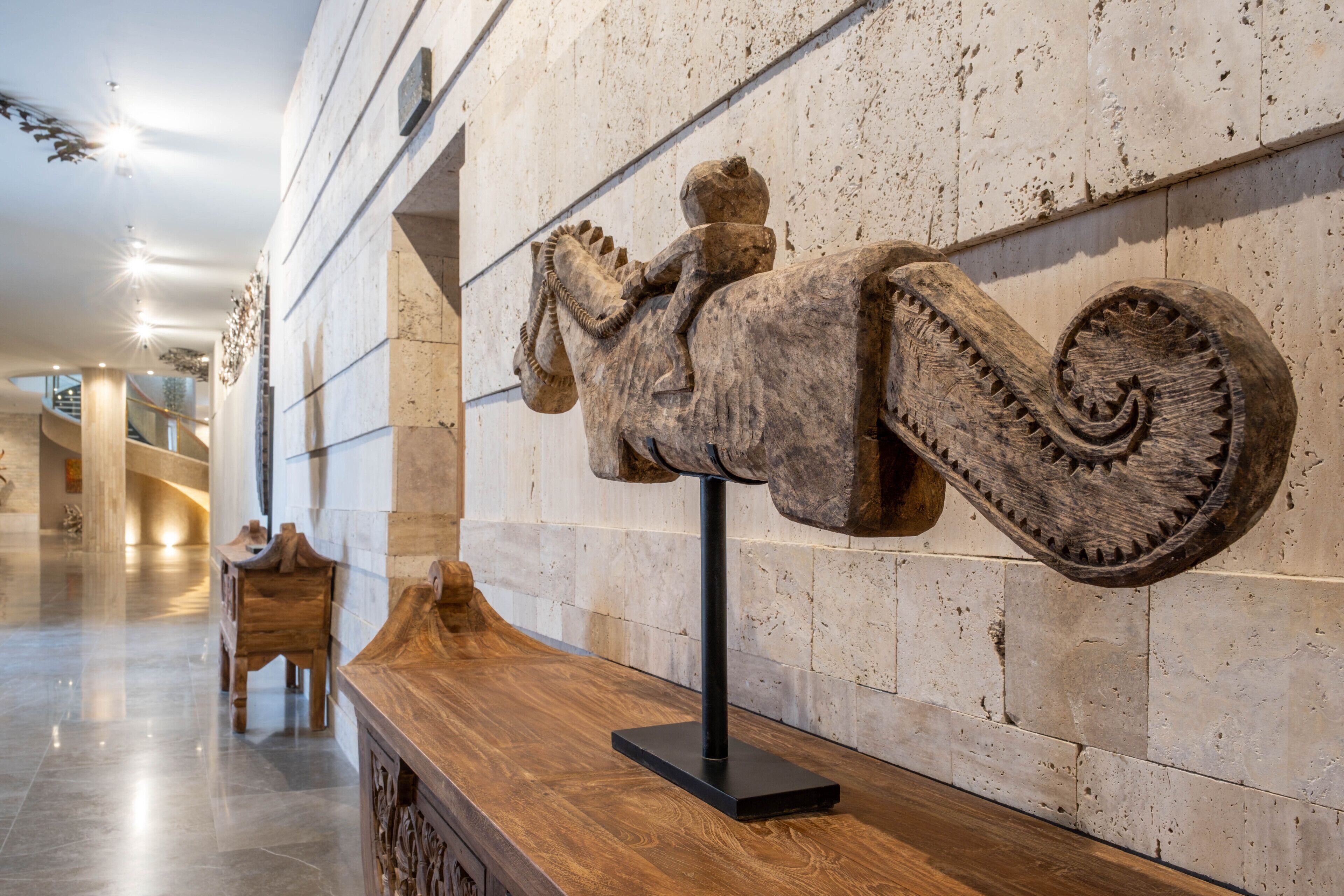 A wooden sculpture of a figure on a horse, mounted on a wall in a lobby with a marble floor and a wooden bench.