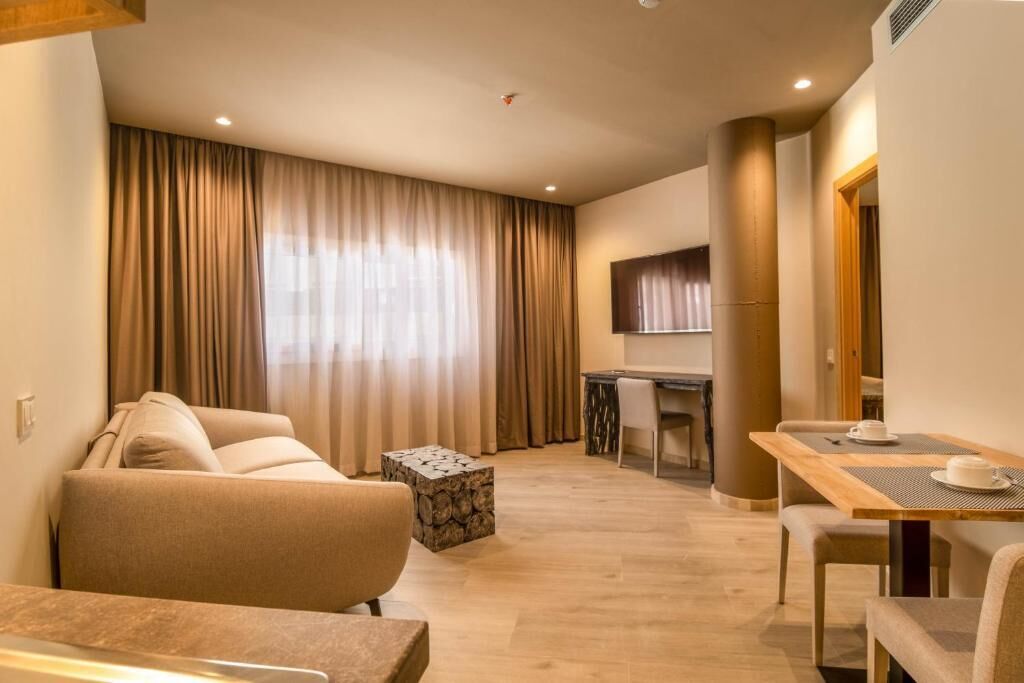Quarto de hotel moderno com sofá, mesa de jantar, televisão e uma janela grande com cortinas.