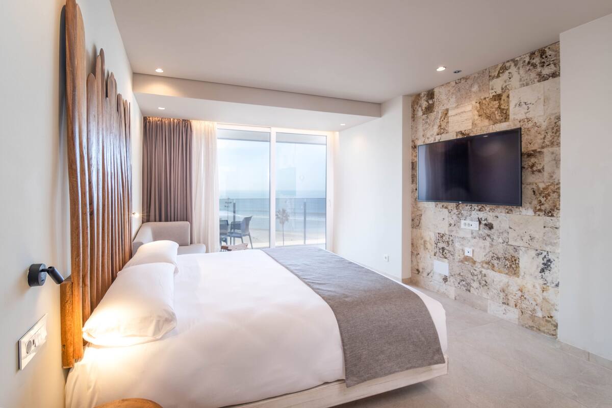A modern hotel room with a large bed, a flat-screen TV mounted on a stone wall, and a view of the ocean through large windows.