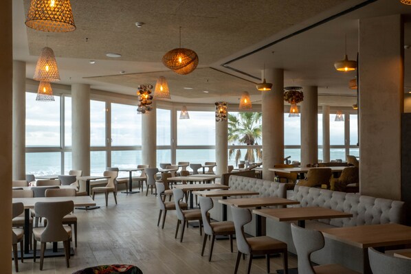 A spacious restaurant with high ceilings, large windows, and a view of the ocean. Tables and chairs are arranged for dining.