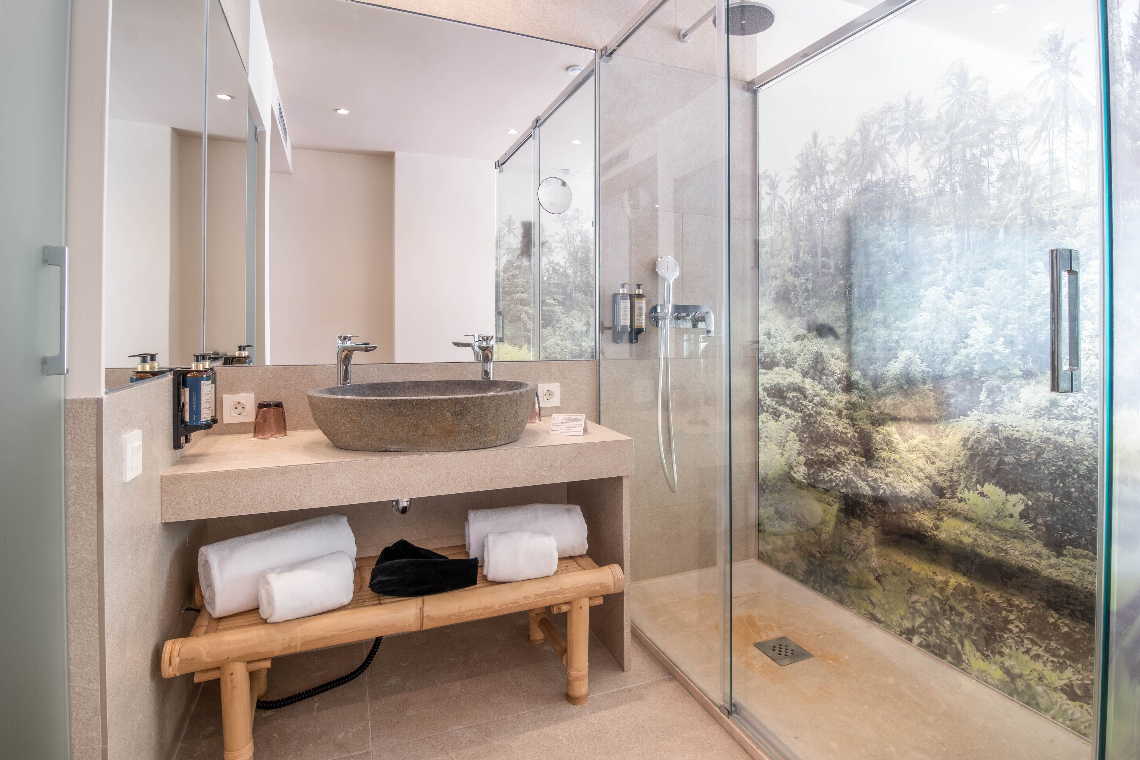 A modern bathroom with a large stone sink, a wooden bench, and a glass shower enclosure with a view of a tropical landscape.