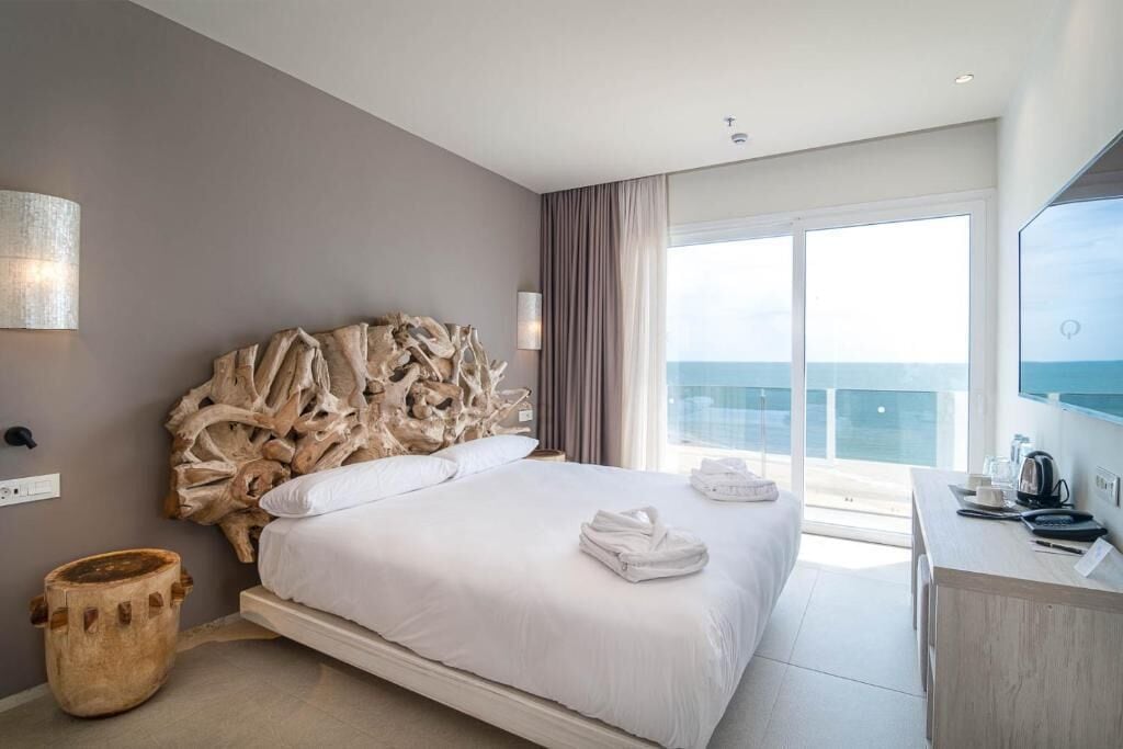 Habitación de hotel moderna con una cama grande, cabecera de madera, un escritorio y un amplio ventanal con vistas al mar.