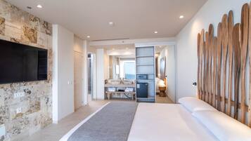 A modern hotel room with a large bed, a flat-screen TV mounted on a textured stone wall, a wooden headboard, and a built-in desk with a mirror and shelves.