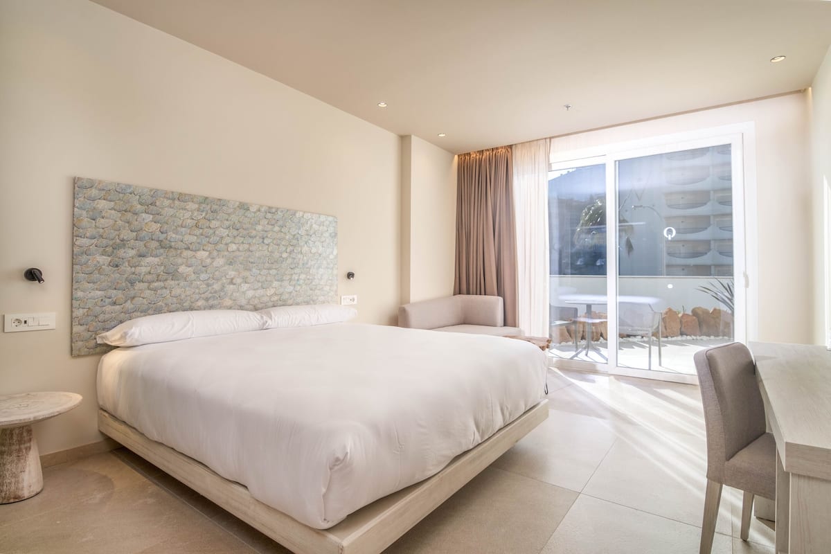 A modern bedroom with a large bed, a chair, a small table, and a balcony with outdoor seating.