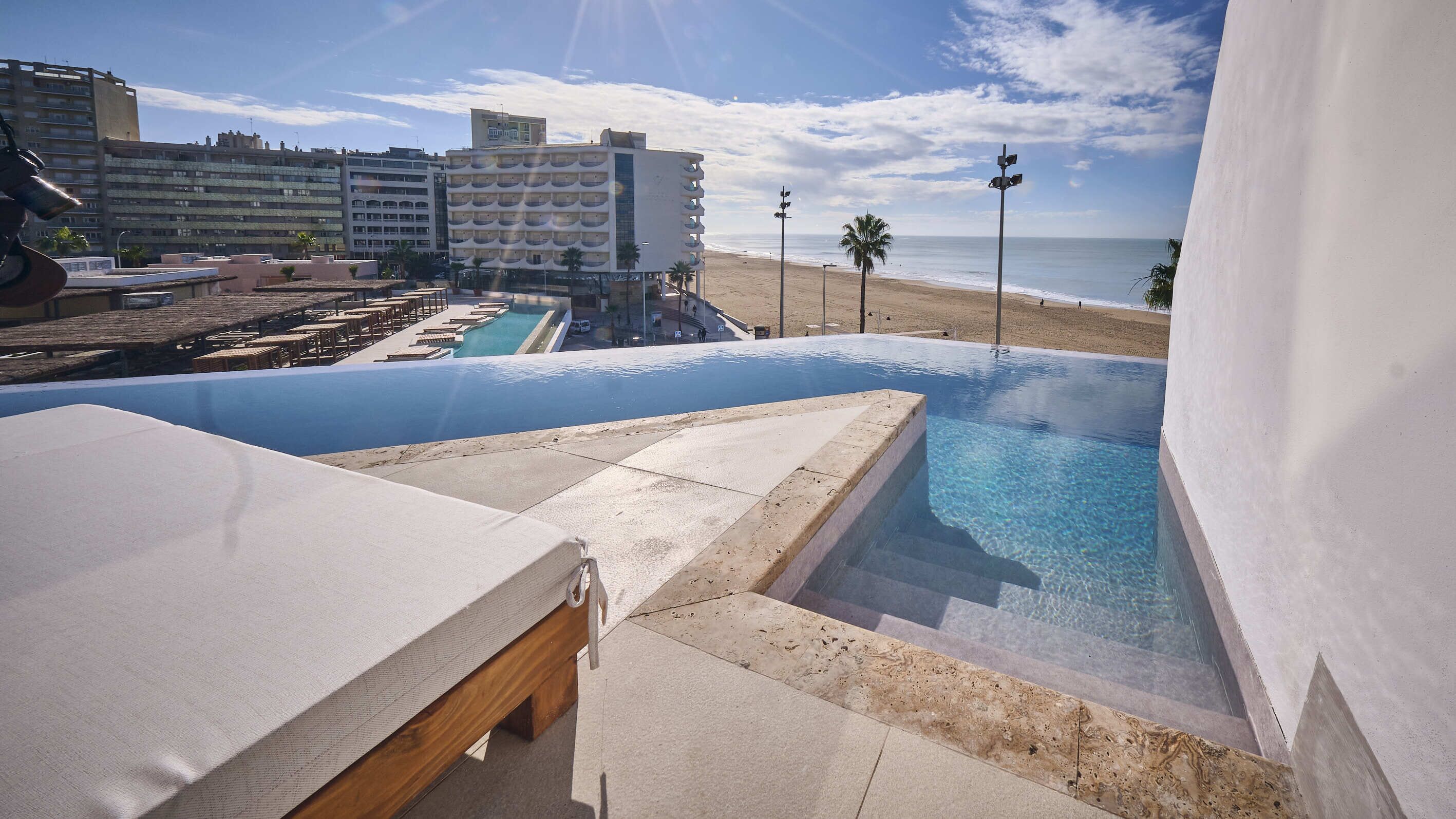 A poolside view with a clear view of the beach and ocean, a lounge area with a white canopy, and a modern building in the background.