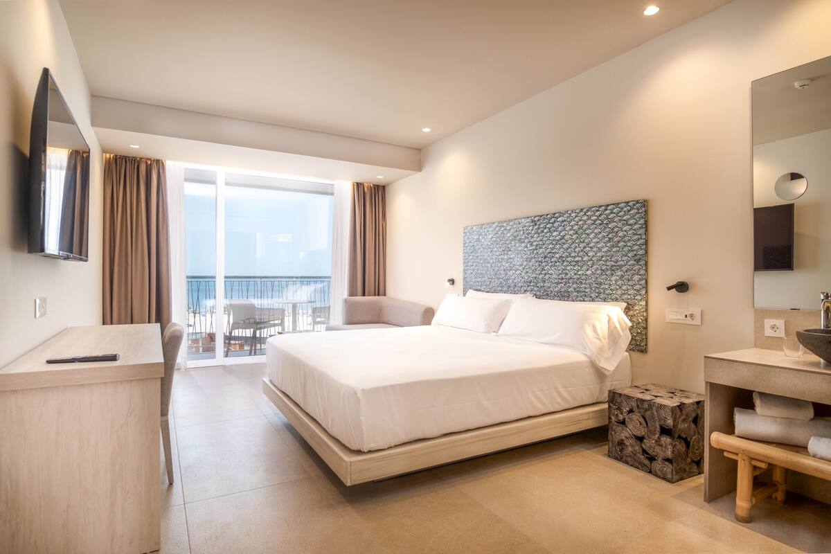 A modern hotel room with a large bed, a desk, a chair, a small table, a bathroom with a sink and mirror, and a balcony with a view.