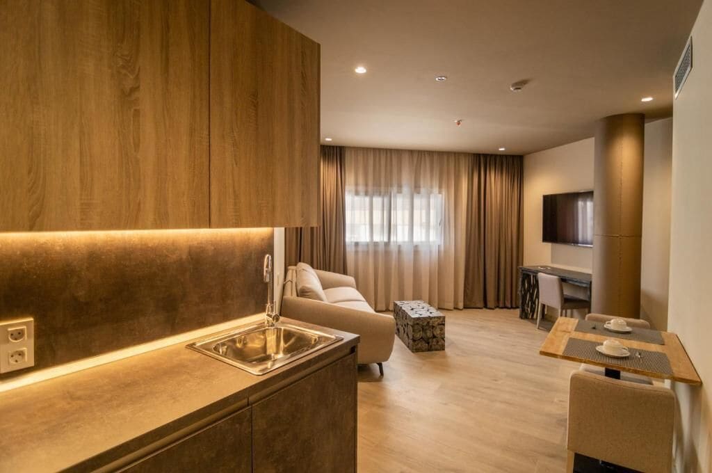 A modern hotel room with a kitchenette, a sofa, a dining area, and a TV.